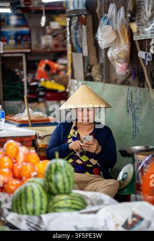 Vendeuse sur un stand de marché de rue vendant des fruits tropicaux frais et défilant sur son smartphone. Banque D'Images