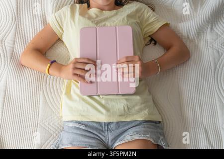 Jeune femme allongée sur un lit, tenant une tablette fermée, se relaxant et interagissant avec des médias numériques Banque D'Images