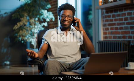 Homme d'affaires afro-américain souriant et heureux passant un appel téléphonique sur mobile et regardant son ordinateur portable le soir dans son bureau à domicile. Homme d'affaires joyeux Banque D'Images
