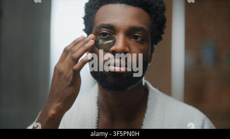 Homme afro-américain avec des patchs oculaires pour hydrater et raffermir la peau des yeux dans la salle de bain à domicile, homme adulte d’origine ethnique profitant des soins personnels, complexion cutanée, anti-rides Banque D'Images