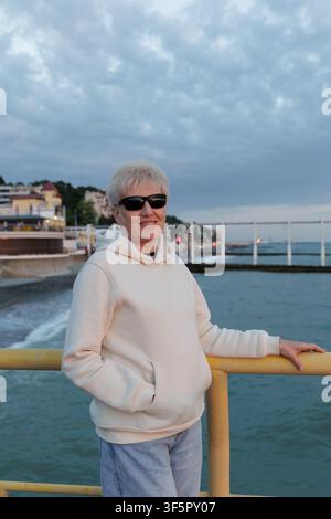 Sénior heureuse aux cheveux blonds courts portant un sweat à capuche décontracté et des lunettes de soleil, se relaxant sur une promenade côtière dans une station balnéaire européenne. Banque D'Images