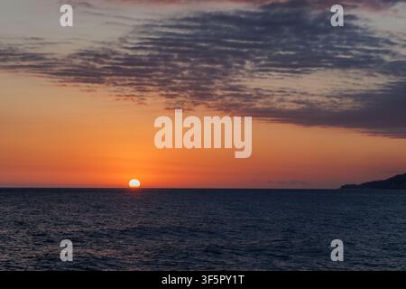 Soleil doré brillant se couchant sur la ligne d'horizon au-dessus de la surface calme de la mer sous un ciel crépusculaire orange. Banque D'Images