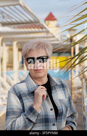 Femme mûre confiante aux cheveux blonds courts portant une chemise à carreaux et des lunettes de soleil, se relaxant dans un complexe côtier moderne pendant l'heure dorée. Banque D'Images