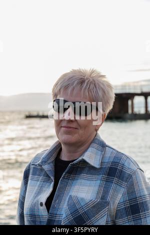 Femme mûre sereine aux cheveux courts portant des lunettes de soleil et une chemise à carreaux se relaxant sur une plage côtière près de l'océan pendant l'heure dorée. Banque D'Images