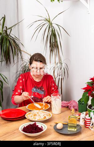 Dans un cadre domestique confortable, une femme mélange des couches de légumes bouillis avec de la mayonnaise pour préparer une salade Olivier. La scène montre des décorations de fête et des plantes festives à proximité Banque D'Images