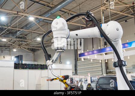 Bras robotique dans un environnement industriel, corps métallique blanc, tuyau noir flexible, plafond élevé, poutres métalliques, panneaux de contrôle, machines, éclairage uniforme et lumineux Banque D'Images