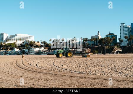 Tracteur d'entretien de plage nettoyant la ligne de côte sablonneuse à Santa Monica, Californie. Motifs lisses dans le sable, palmiers, bâtiments modernes Banque D'Images