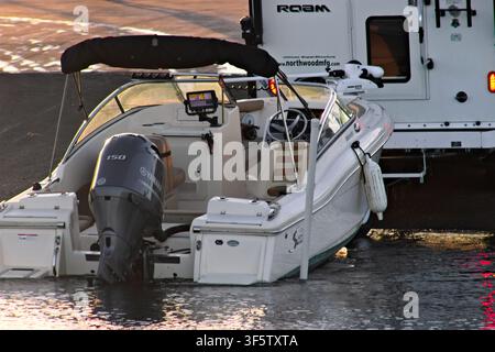 Bateau à moteur blanc avec moteur hors-bord noir flotte sur l'eau calme. Un auvent noir recouvre le cockpit. Une remorque se trouve derrière le bateau sur la rive. Teintes du coucher de soleil li Banque D'Images