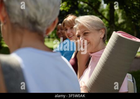 Sénior heureuse tenant un tapis de yoga et discutant avec des amis dans le parc Banque D'Images