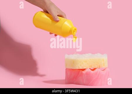 Main versant du savon à vaisselle d'une bouteille jaune sur une pile d'éponges avec de la mousse sur un fond rose Banque D'Images