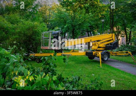 Plate-forme de travail aérienne jaune avec panier à bras étendu en métal et contre-poids sur pelouse verte à côté d'un chemin pavé, entourée de buissons et d'arbres denses Banque D'Images