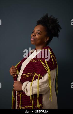 Jeune femme afro-américaine posant dans un foulard tricoté à la main, regardant au loin. Image idéale pour les campagnes de mode hivernale, le marketing artisanal et les éditoriaux lifestyle, verticale Banque D'Images