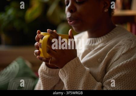 Jeune femme afro-américaine tenant une tasse jaune, soufflant sur une boisson chaude, pratiquant la pleine conscience. Image adaptée aux campagnes de bien-être mental et au marketing de style de vie, plan rapproché Banque D'Images