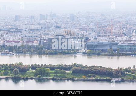 Vue aérienne du paysage urbain de Vienne se déployant derrière le Danube et la verdure luxuriante de l'île du Danube sous un ciel brumeux. Bâtiments modernes et éloignés Banque D'Images