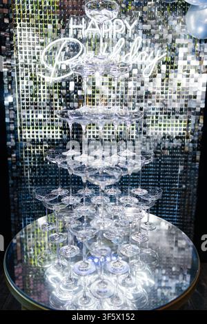 Pyramide de verres à champagne vides disposés sur une table ronde en verre devant un panneau néon lumineux « joyeux anniversaire » et un fond à paillettes brillantes lors d'une fête Banque D'Images