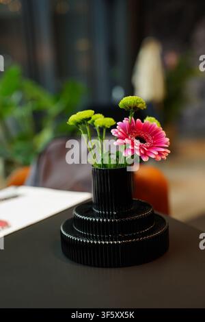 Petit bouquet comprenant une gerbera rose et des fleurettes vertes dans un vase noir à côtes unique à étages, posé sur une table sombre, éclairage intérieur doux Banque D'Images