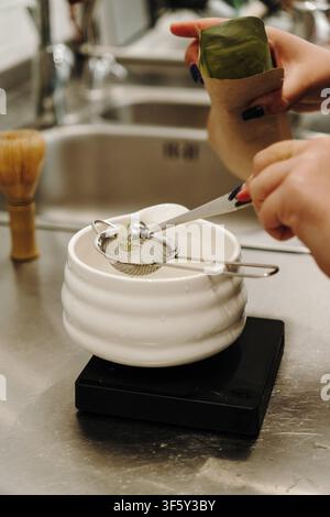Mains mesurant de la poudre de matcha premium à travers une passoire fine dans un bol en céramique. Préparation traditionnelle du thé japonais avec des outils de précision sur un Banque D'Images