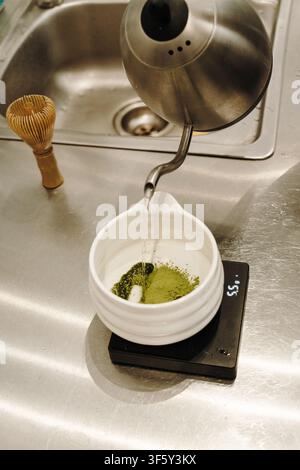 De l'eau chaude est versée sur de la poudre de matcha tamisée dans un bol en céramique posé sur une balance numérique. Préparation traditionnelle du thé japonais en cours. Banque D'Images