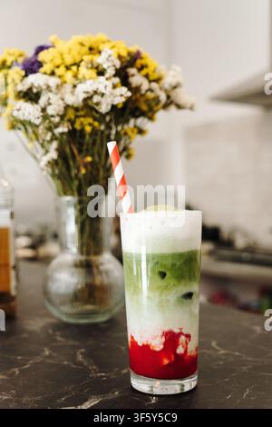 Latte matcha aux fraises en couches garni d'une mousse crémeuse et servi avec une paille en papier rayée. Boisson spéciale rafraîchissante en gros plan. Banque D'Images