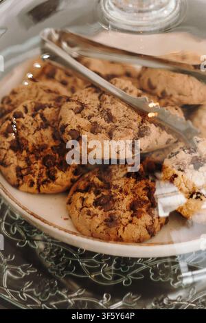 Des cookies aux pépites de chocolat frais exposés sous un dôme en verre avec une pince à servir dans un café moderne. Pâtisseries maison présentées pour les clients. Banque D'Images