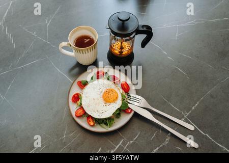 Petit-déjeuner sain comprenant un toast à l'avocat avec un œuf au plat, du thé chaud et une French press. Composition épurée avec lumière naturelle et espace de copie. Banque D'Images