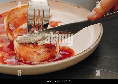 Close up de crêpes moelleuses au fromage blanc coupées avec un couteau et une fourchette sur une assiette avec une sauce aux fruits rouges. Dessert de petit-déjeuner fait maison servi dans un moderne Banque D'Images