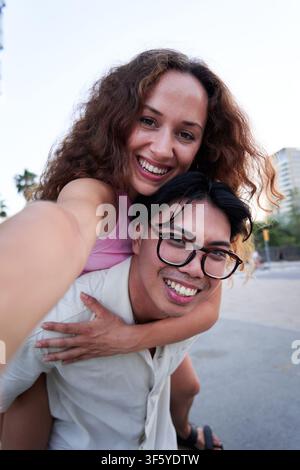 Selfie de jeune couple regardant la caméra tandis que le petit ami donne piggyback à sa petite amie. Banque D'Images