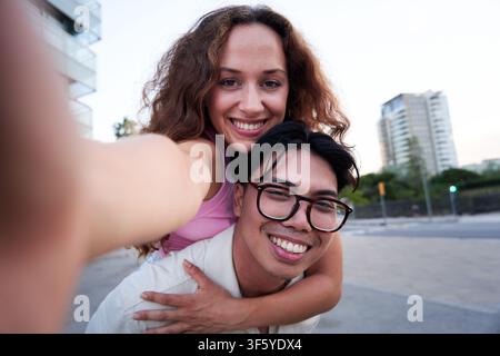 Selfie de jeune couple regardant la caméra tandis que le petit ami donne piggyback à sa petite amie. Banque D'Images