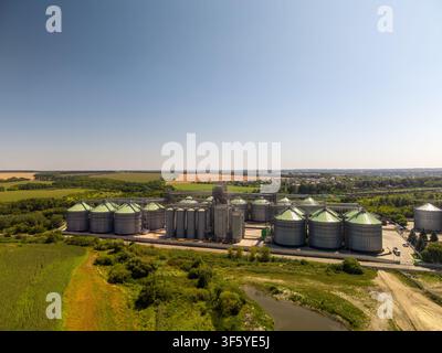 Vue panoramique en contre-plongée d'un terminal industriel de stockage de céréales à grande échelle avec des silos métalliques entourés de campagne verdoyante. Banque D'Images