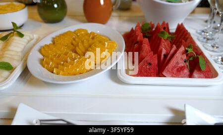 Tranches de pastèque rouge vif et de mangue dorée disposées soigneusement sur des assiettes en céramique blanche avec des feuilles de menthe fraîche. Un éclairage naturel vif met en valeur le te juteux Banque D'Images