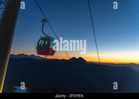 Une vue aérienne époustouflante capture une voiture gondole rouge suspendue à un téléphérique, glissant gracieusement devant un arrière-plan de coucher de soleil à couper le souffle. Silhouettes de montagnes Banque D'Images