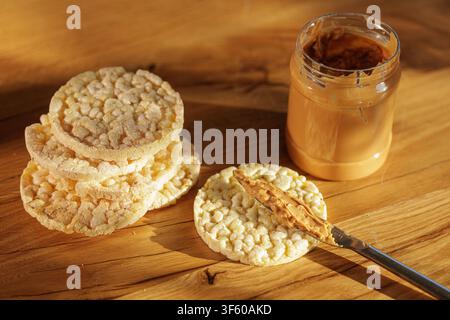 Pot de beurre de cacahuète avec couteau étalant la pâte sur des gâteaux de riz croustillants sur une table en bois, idéal pour les collations saines, les promotions de régime ou de nutrition. Banque D'Images