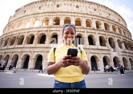 Femme noire joyeuse regardant l'appareil photo tout en tenant un téléphone portable avec le Colisée en arrière-plan. Banque D'Images