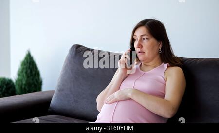 Femme enceinte ayant une conversation inquiète sur son smartphone à la maison Banque D'Images