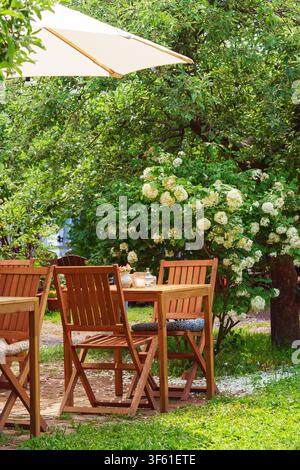 Table et chaises en bois dans une terrasse de jardin accueillante, café en plein air, restaurant entouré d'une végétation luxuriante et d'arbustes en fleurs. Espace extérieur paisible d Banque D'Images
