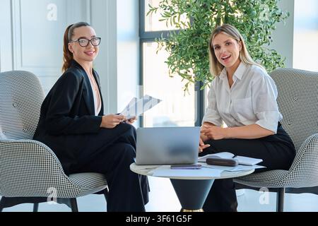 Deux femmes d'affaires prospères souriant à l'appareil lors d'une réunion dans un bureau moderne Banque D'Images
