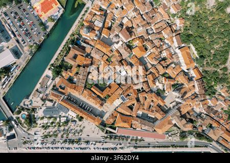 Photographie par drone montrant l’agencement médiéval dense de la vieille ville de Kotor au Monténégro, avec des toits en terre cuite, des rues étroites et sinueuses et des remparts environnants, bordés par un canal et des zones de stationnement. Banque D'Images