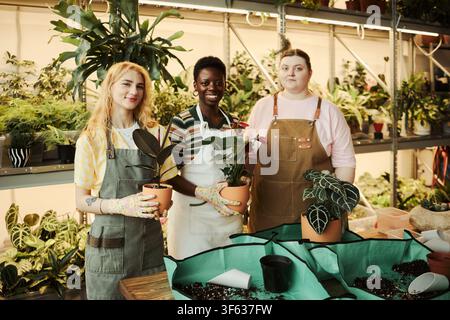 Portrait de jeunes fleuristes diversifiées souriant en tenant des plantes en pot dans une serre. Image adaptée au marketing des petites entreprises et à la publicité pour les magasins de plantes locaux Banque D'Images