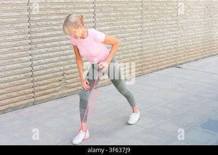 Une personne s'étirant avec des bandes de résistance dans un cadre urbain en plein air, concentrée et active, vue de côté, haut rose et leggings verts, pavé en béton Banque D'Images
