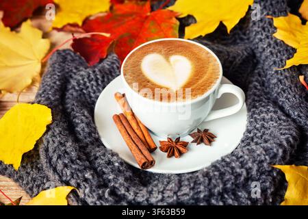 Nature morte automnale douillette mettant en scène une tasse de cappuccino crémeux avec un cœur en latte art, des bâtons de cannelle et des feuilles d'automne colorées sur une couverture tricotée. Caché Banque D'Images