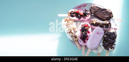 Ronde colorée de bâtonnets glacés maison au yaourt et au lait. Vanille et chocolat décorés de noix et de baies surgelées sur un fond bleu vif. Parfait pour une fête d'anniversaire d'enfants en été. Banque D'Images