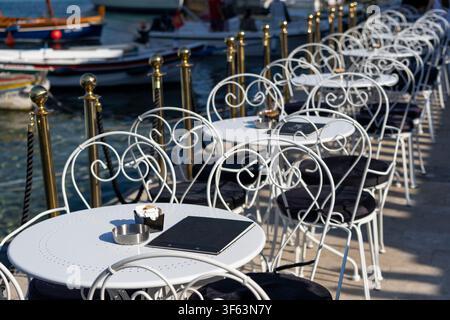 Range de tables et chaises vides en fer forgé blanc sur la terrasse extérieure d'un café au bord de l'eau calme, lumière vive, rampes métalliques décoratives, b amarré Banque D'Images