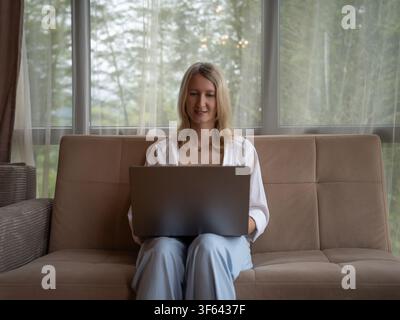 Femme blonde souriante, se concentrant sur son ordinateur portable tout en étant confortablement assise sur un canapé moderne près d'une fenêtre, profitant du travail à distance et de la communication en ligne Banque D'Images