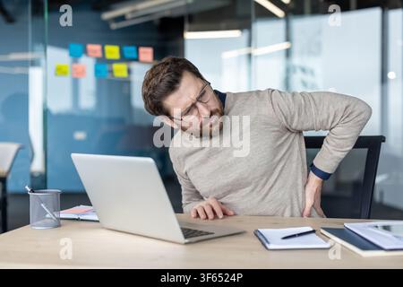 Homme ressentant une douleur aiguë au bas du dos en travaillant sur un ordinateur portable dans un bureau moderne, mettant en évidence l'impact du travail sédentaire et d'une mauvaise ergonomie sur la santé et le bien-être Banque D'Images