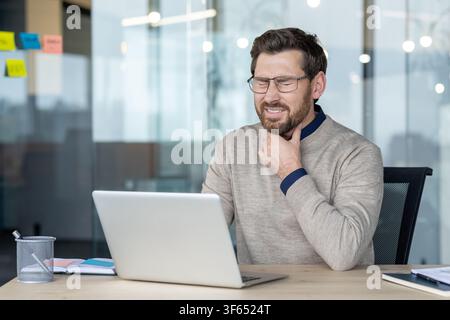 Homme ressentant une douleur intense due à un mal de gorge et à la maladie, touchant son cou alors qu'il est assis à un bureau avec un ordinateur portable, reflétant les défis du travail en état de maladie Banque D'Images