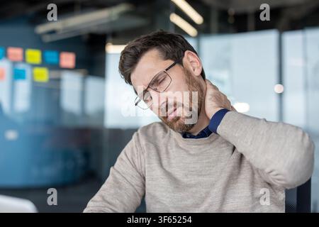 Homme d'affaires stressé souffrant d'un cou raide et de maux de tête, se frottant la main sur sa nuque douloureuse, ressentant un malaise et une tension lors du travail dans un environnement de bureau moderne Banque D'Images