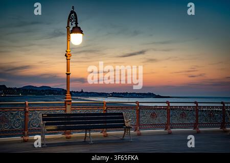 Un lampadaire orné de style victorien allumé à côté d'un banc vide sur le Victoria Pier au coucher du soleil sur la plage de Colwyn Bay, dans le nord du Pays de Galles, Royaume-Uni. Banque D'Images