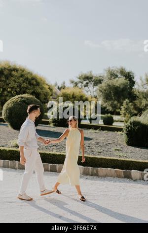 Un couple profite d'une promenade tranquille, main dans la main, à travers un jardin vibrant. Le soleil brille fortement, illuminant leurs moments joyeux. Banque D'Images