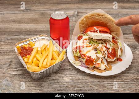 Main tenant un sandwich kebab chargé à côté de frites avec des sauces et une canette de soda rouge sur une table en bois Banque D'Images