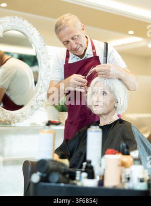 Le maître expérimenté de l'homme senior fait effectue la coupe de cheveux pour la femme mature avec des ciseaux Banque D'Images